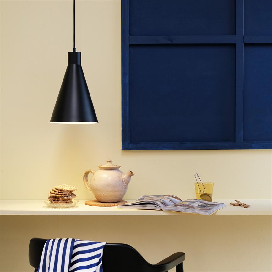 Black pendant lamp casts warm light over a ceramic teapot, stack of biscuits, open book and glass with tea strainer on a light shelf in front of a dark blue wall panel.