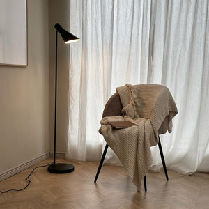 An upholstered chair covered in a beige woven rug with a book on the seat; a lit black floor lamp shines in front of white curtains in a bright, minimalist room.