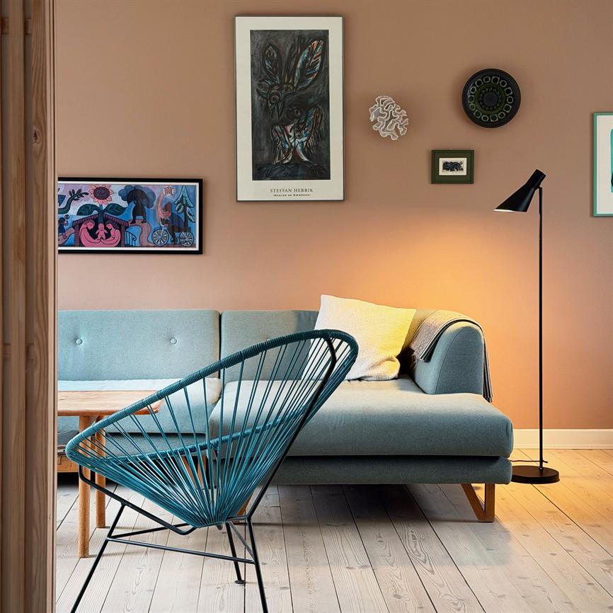 An upholstered chair covered in a beige woven rug with a book on the seat; a lit black floor lamp shines in front of white curtains in a bright, minimalist room.