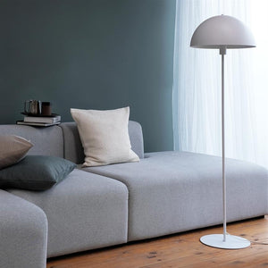 A light gray modular sofa is furnished with cushions and books; a white floor lamp stands by a bright, curtained window in a quiet, dark green room with a wooden floor.