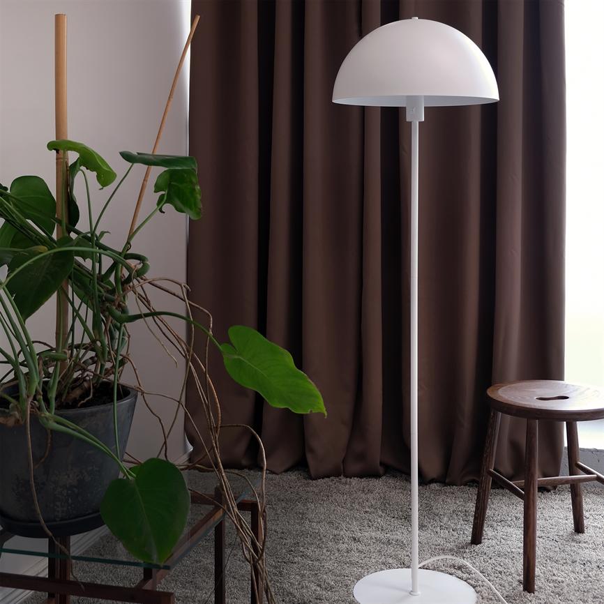 White hemispherical floor lamp stands in front of dark brown curtains on a gray rug, flanked by a large potted plant with long vines and a wooden stool by the window.