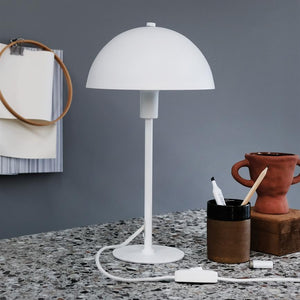 A white table lamp stands on a terrazzo table; its dome sits on a slender cane. The background is a gray wall with papers and an embroidery frame; to the right pen holder and terracotta pot.