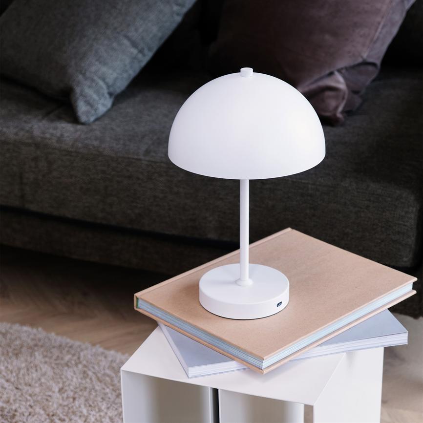 A white, half-round table lamp is switched off on a stack of folders on a small white side table; dark gray sofa and cushions in the background, light carpet and wooden floor below.