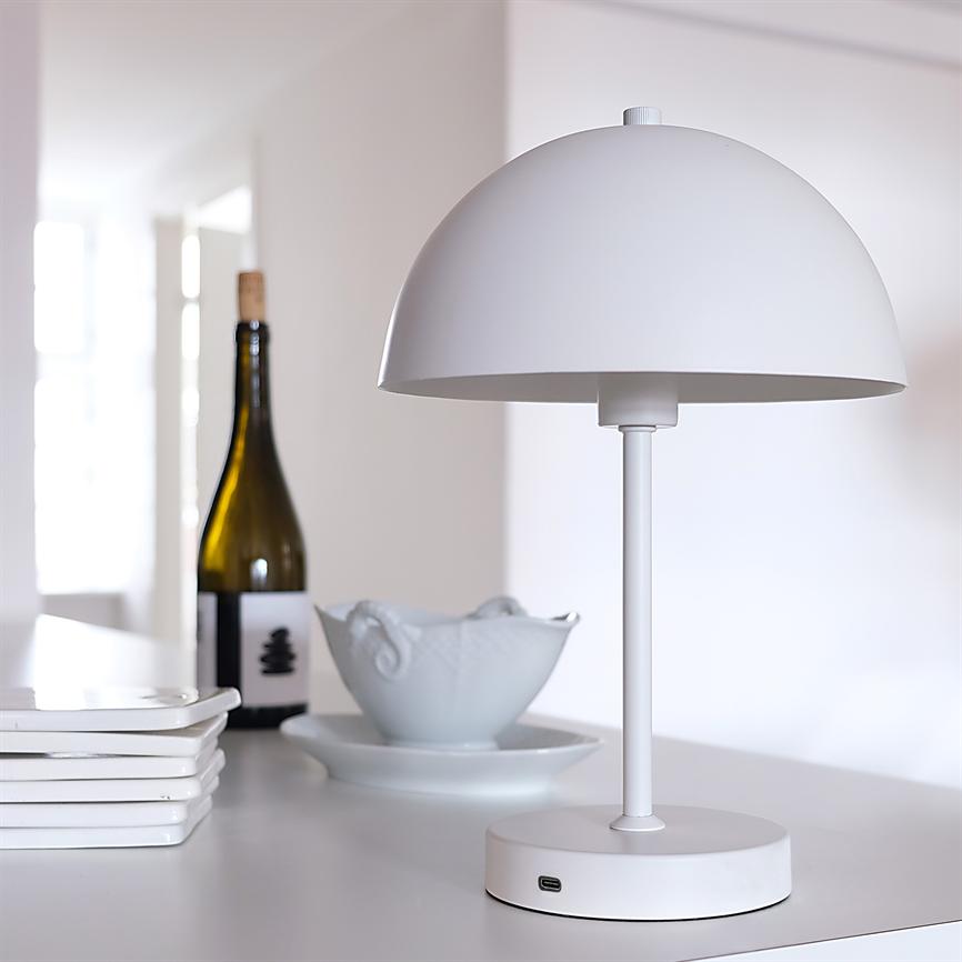 A white, hemispherical table lamp stands on a white table top with a visible USB port; it is flanked by a wine bottle, a ceramic bowl and a stack of white plates in a bright, minimalist space.
