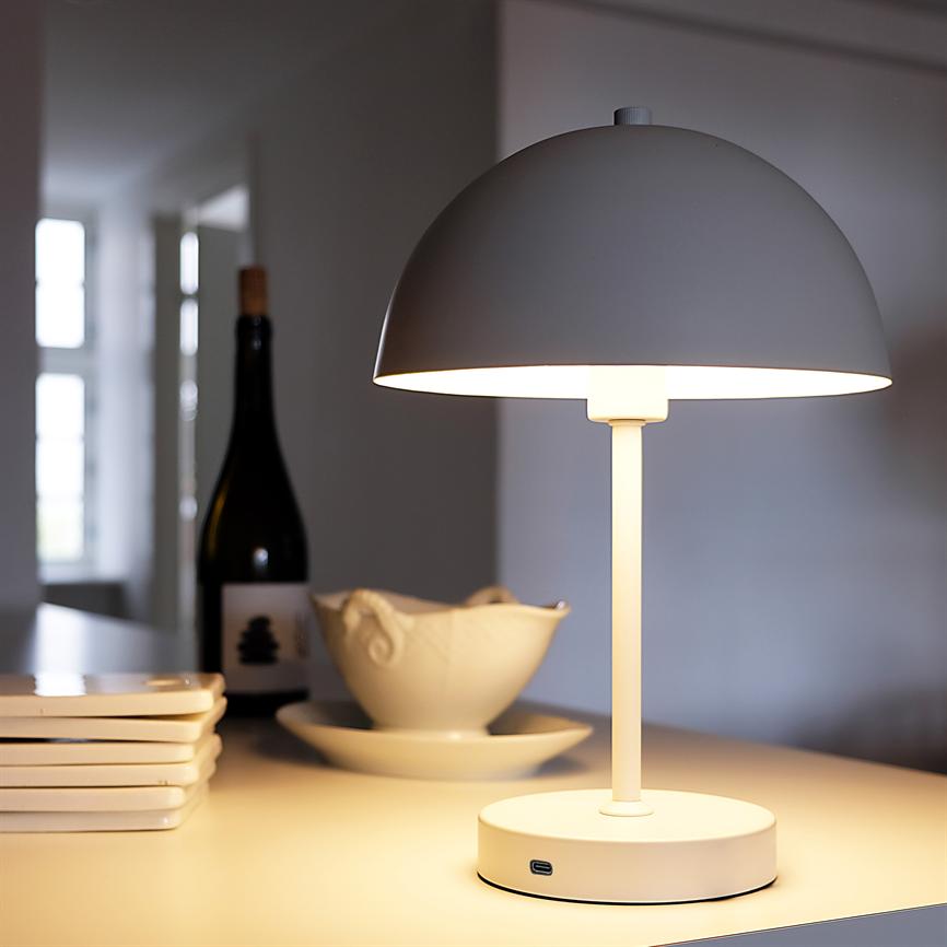 Table lamp lights up on the table; surrounded by a stack of plates, a sauce pot and a bottle of wine in a bright, calm kitchen environment.