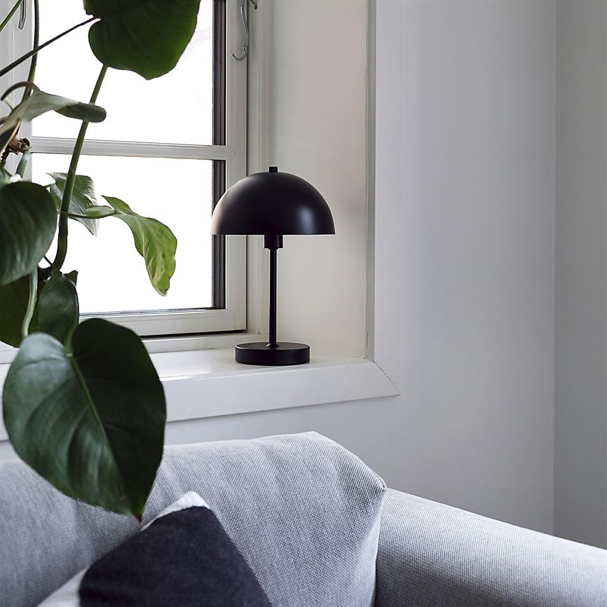Black table lamp is switched off on the windowsill; green plant leaves hang against the window, and a light gray sofa with a dark cushion is seen in a bright, minimalist room.