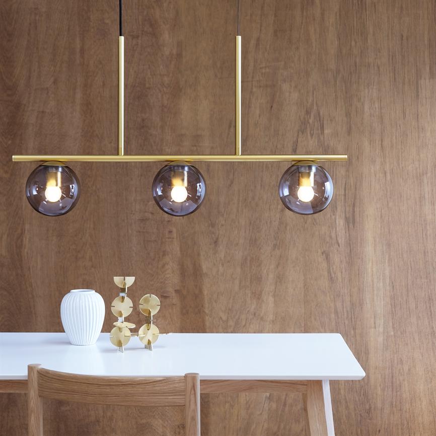Triple pendant lamp with three smoky glass balls hangs above a white table and casts warm light; a wooden chair, white vase and gold colored decoration stand in front of a wood paneled wall.
