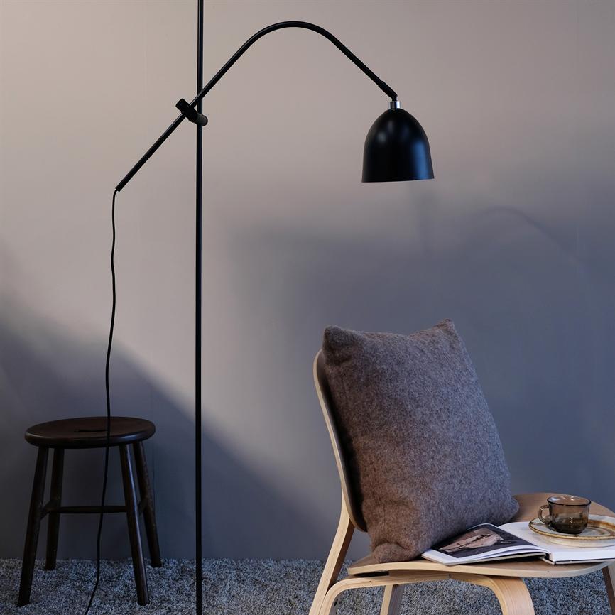 Arc-shaped black floor lamp hangs above a wooden chair with gray cushion; the chair is on a gray rug by a footstool, with an open book and a glass cup on the seat.