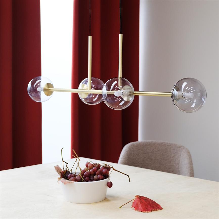 A modern gold-colored ceiling lamp with four clear glass domes hangs above a bright dining table; a white bowl with red grapes is on the table in front of deep red curtains and a soft chair.