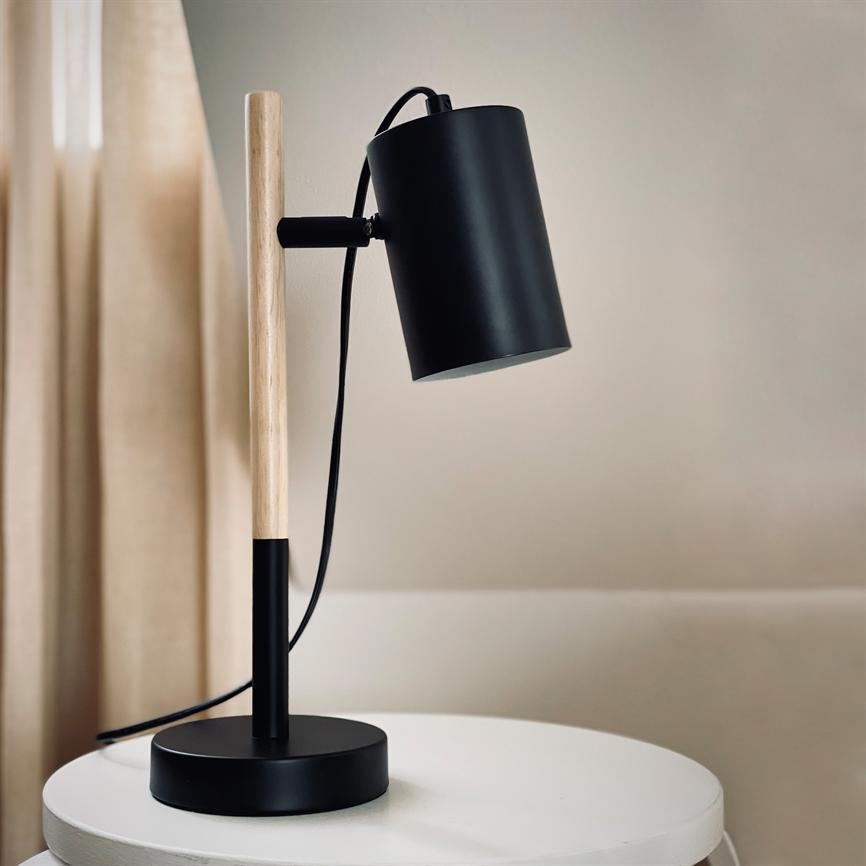 A black and wooden table lamp stands on a round white table; the lamp head is angled downwards, the cord is hanging; background: beige wall and light curtains.