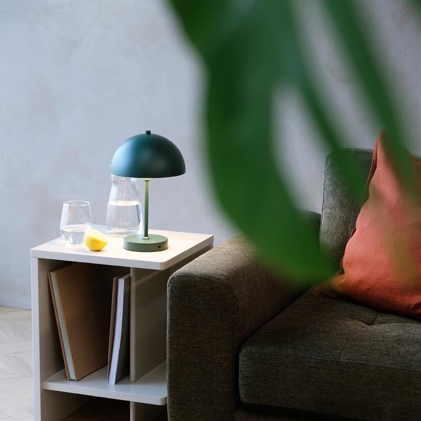 Green table lamp casts soft light over a white side table with jug, glass and lemon, placed by a gray sofa with rust-coloured cushion; a green plant leaf silhouette in the foreground.