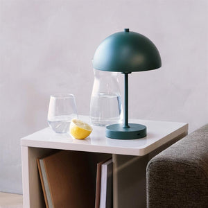 A green table lamp stands on a white side table; a glass carafe, a glass of water and half a lemon lie beside it; the table is next to a gray sofa in front of a light wall.