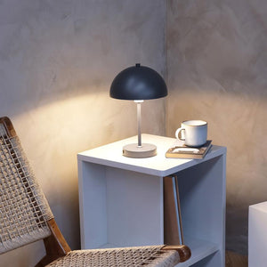 A black dome lamp glows softly on a white bedside table, next to a cup and books, in a cozy corner with a wicker chair and a raw, textured wall.