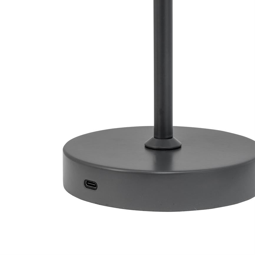 The lamp's round, dark metal base stands on a white background; a slim post rises from the center, and a USB-C port is built into the side of the foot.