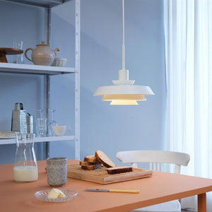 A white one pendant shines warmly above a kitchen table, where a cutting board with slices of bread stands with a jug of milk, butter, a mug, a shelf with crockery and a white chair by the curtain.