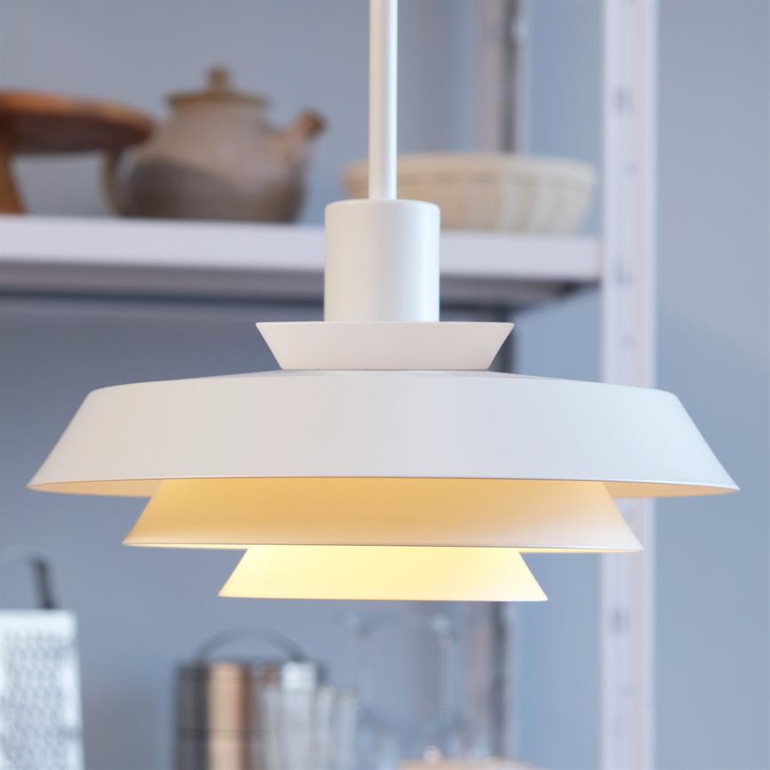 Pendant lamp in white, layered design hangs and emits warm, soft light in front of a blurred kitchen shelf with jars and utensils.