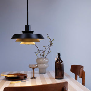 Black tiered ceiling lamp casts warm light over a striped set table with plates, cocktail glasses, brown bottle and vase with branches in a bright, simple dining room.