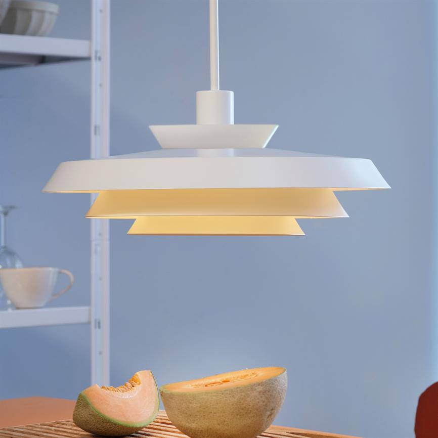 Hanging white pendant emits warm, yellow lighting over a table. On the table, halved cantaloupe; in the background shelf with cup and glass, muted blue wall.
