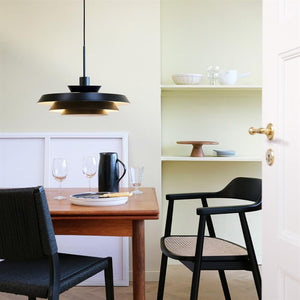 A black pendant lamp hangs and casts warm light over a wooden dining table; glasses, a black jug and plate lie on the table, surrounded by black chairs and pale yellow shelves with ceramics.