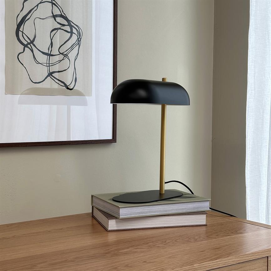 A black and gold table lamp is switched off on two stacked books on a wooden cabinet, in front of a light wall with a framed abstract drawing and a white, transparent curtain.