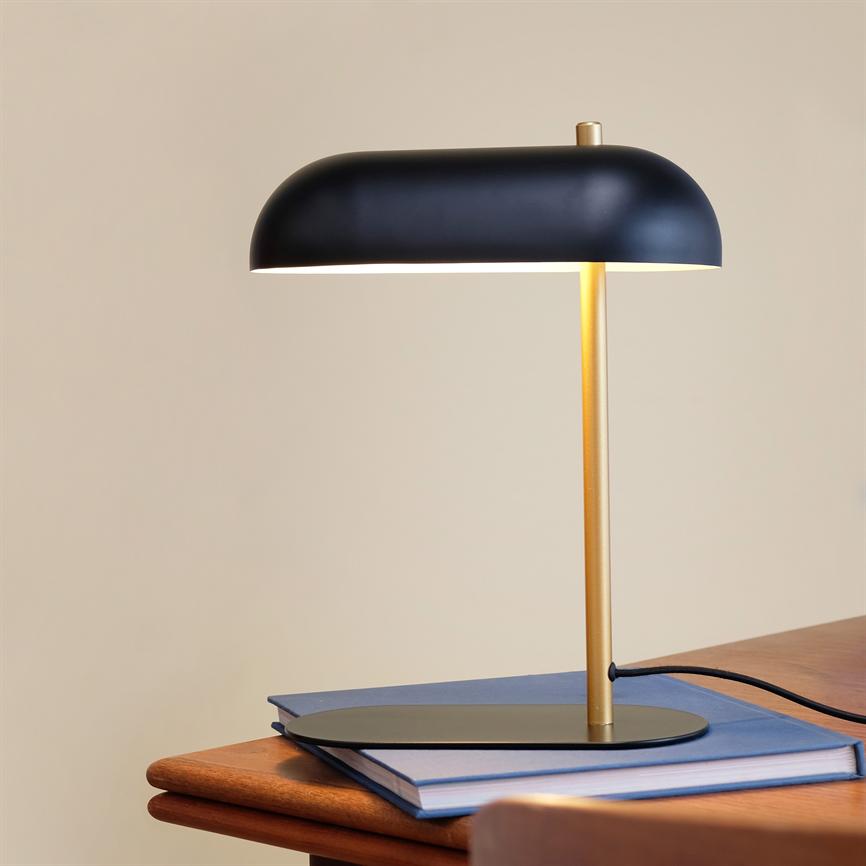 A black table lamp is lit and casts warm, subdued light on two books; placed on a wooden table in front of a neutral beige wall.