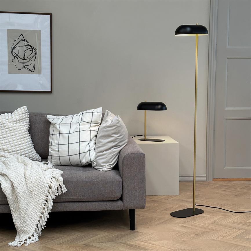 Gray sofa reclines with checked and textured pillows and knitted rug; a black and gold floor lamp and table lamp stand next to it under a framed abstract picture in a bright, modern living room.