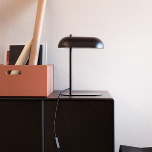 Black table lamp stands on a dark cabinet, cord hanging down; next to a terracotta storage case with scrolls and books, in front of a light, smooth wall.