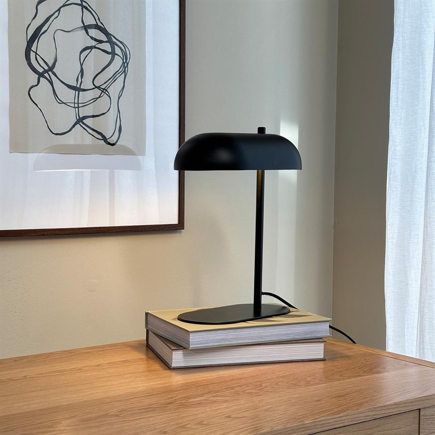 Black table lamp stands on two books on a wooden table, by a wall with framed abstract art and a bright curtain, with a visible cord.