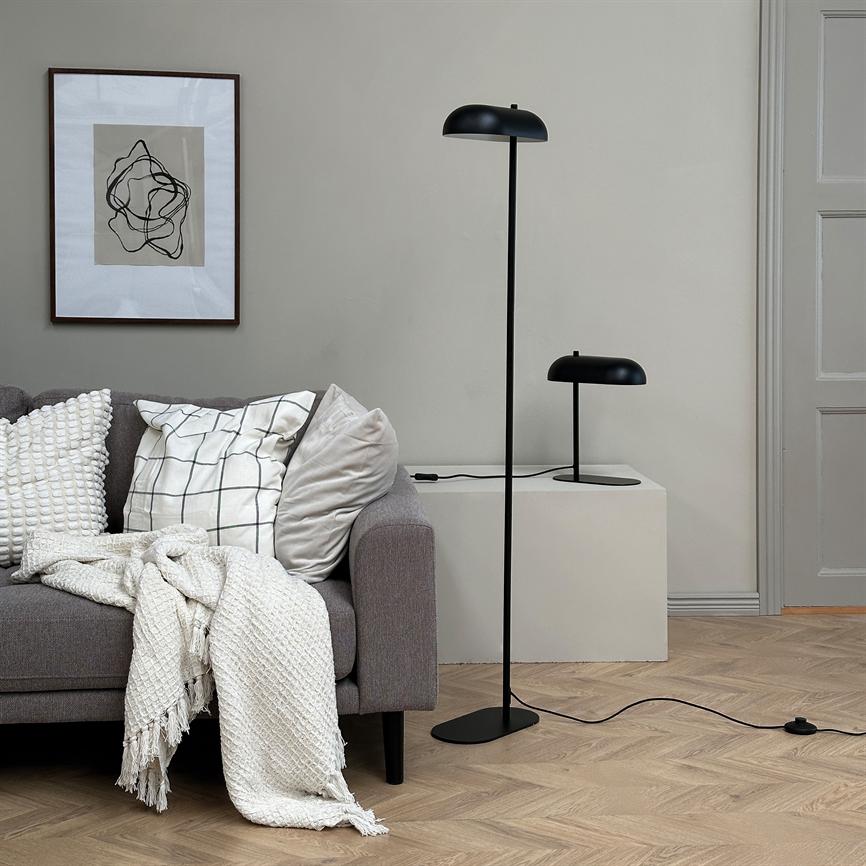 Black floor lamp stands by a gray sofa with checkered cushion and white rug, in front of a light wall with abstract framed art and a black table lamp on a white table.