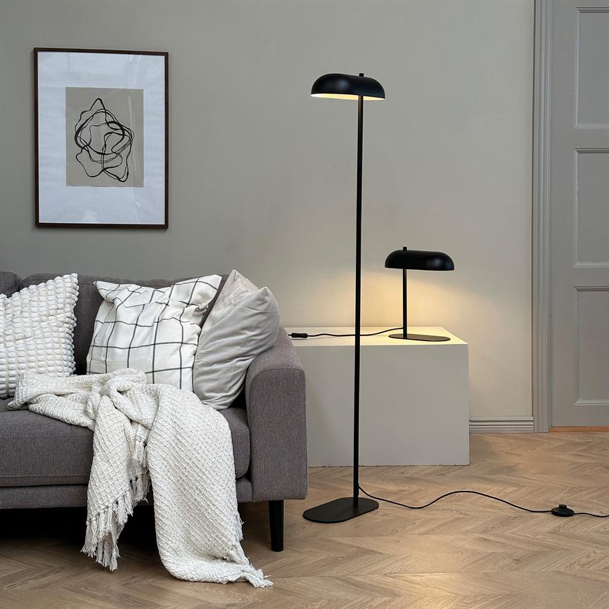 A floor lamp is on and casts warm light over a gray sofa with cushions and a white knitted rug, in front of a white sideboard with a smaller table lamp and framed abstract art.