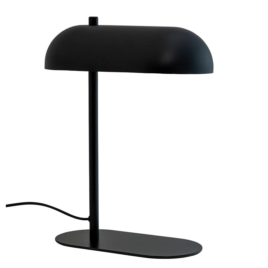 Black table lamp stands on an oval base; slender vertical stand carries a rounded shade, cord runs from foot; placed against a uniform white background.