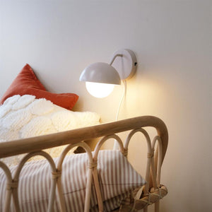A round white wall lamp shines softly above the edge of the bed; a rattan bed with curved slats, a striped pillow, a white plush duvet and an orange throw pillow in a bright bedroom.
