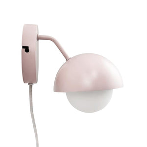 Wall lamp in powder pink hangs mounted on a round wall bracket; a hemispherical shade covers a white opal glass ball, with curved arm and fabric-wrapped cord against a white background.