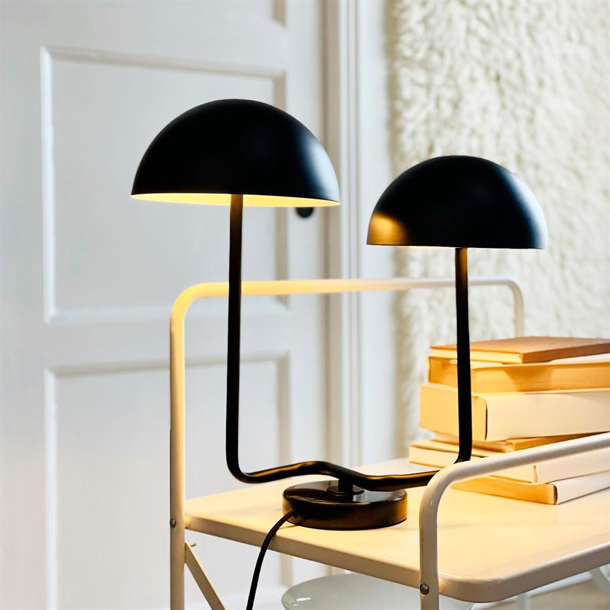 Two black dome lamps stand on a white metal rolling table, casting warm light over a stack of books, in front of a white door and a textured, light wall.