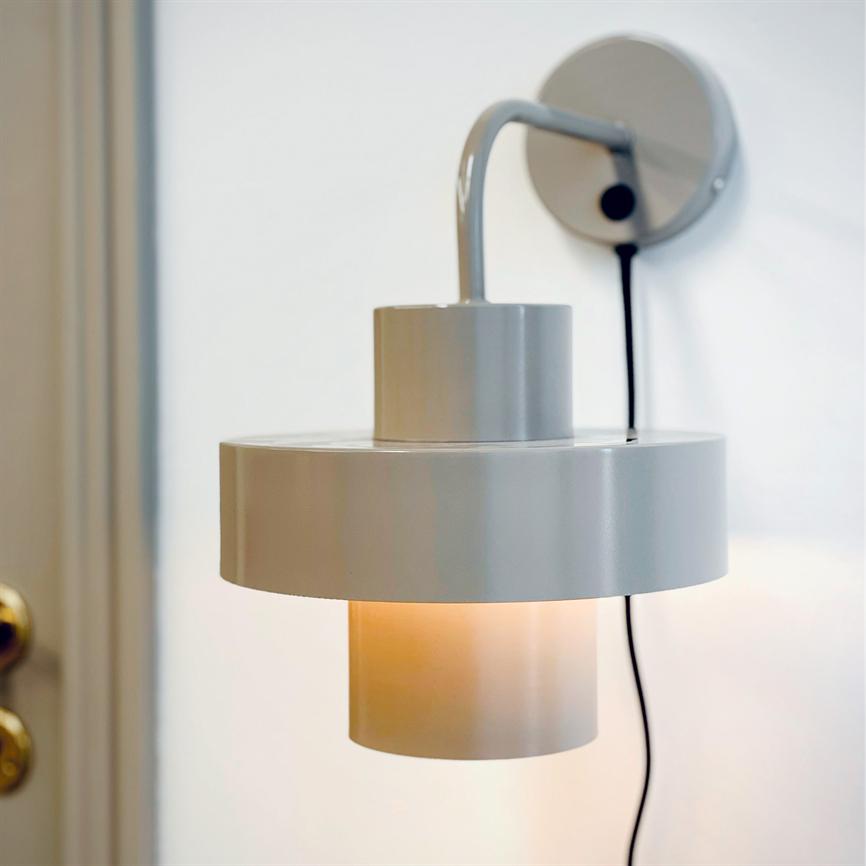 White, round wall lamp shines softly downwards; it is mounted on a white wall with a visible black cord, near a doorknob.