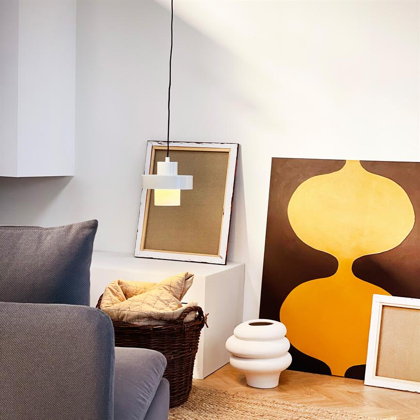 White pendant lamp hangs above a white sideboard; in front is a basket of cushions, a white vase and a large tan abstract painting next to a gray sofa in a bright, minimalist room.
