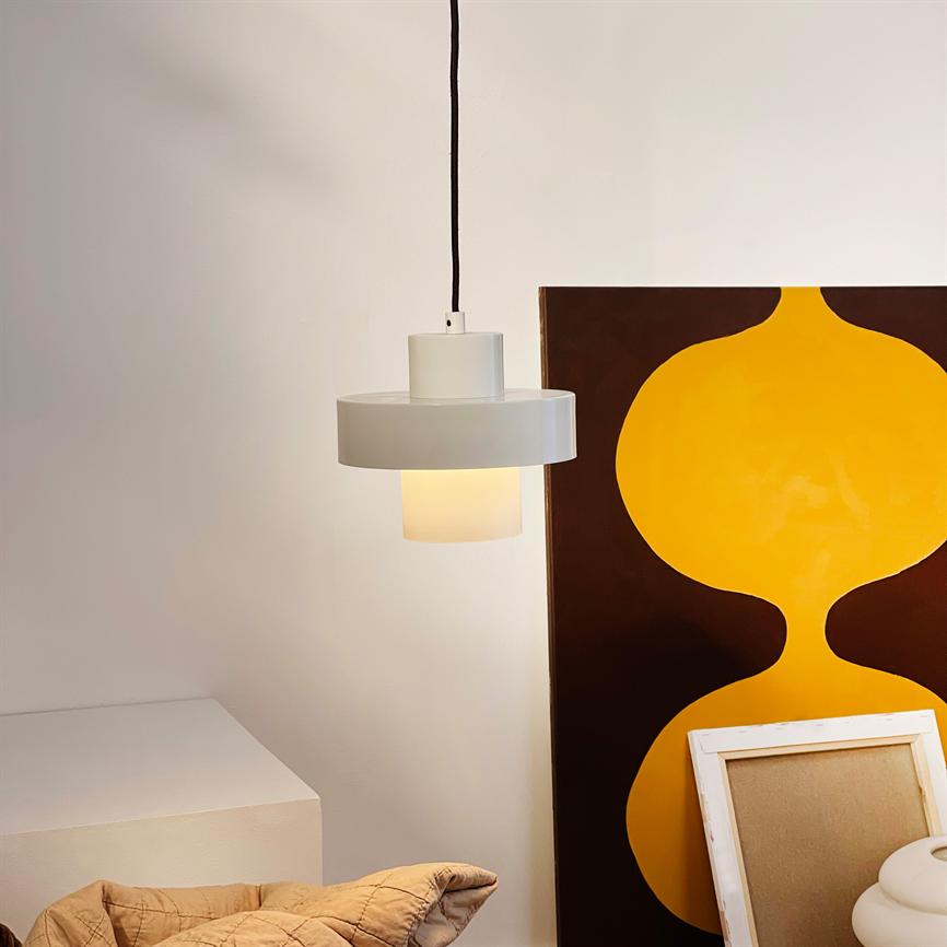 A white hanging lamp hangs from a black cord and emits soft, warm light; it floats in front of a large painting with yellow abstract form, by stacks of frames and a beige carpet.