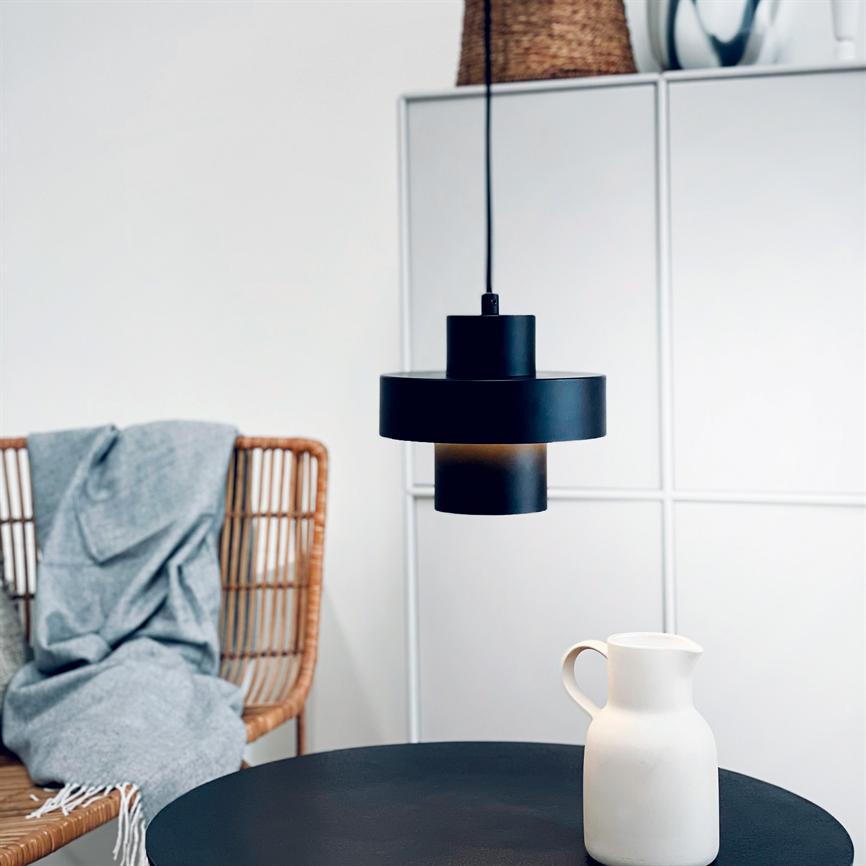 Black hanging lamp hangs above a round black table; a white ceramic jug is on the table; a rattan chair with a gray carpet and white cabinets can be seen in the background.