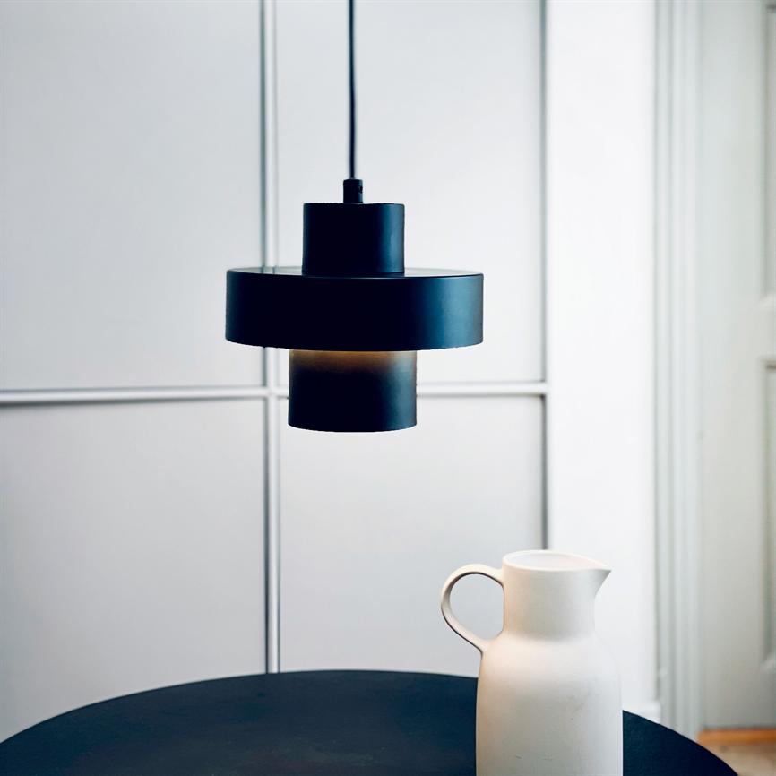Black pendant-lamp hangs and casts soft light over a round dark table, where a white ceramic jug stands; minimalist, bright room with divided wall panels in the background.