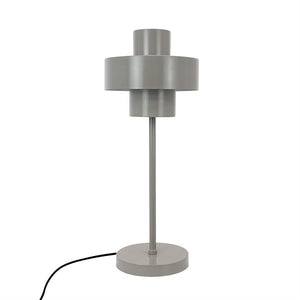 Grå bordlampe står lodret på en rund fod; tynd central stang bærer lagdelt cylindrisk skærm; mat metalfinish; sort ledning ligger bøjet til venstre på hvid baggrund.