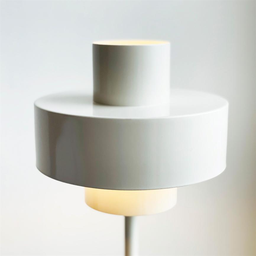 Modern white cylindrical lamp, two stacked shades, emits soft warm light at the bottom; stands on a slender pole in front of a light, neutral wall in a minimalist room.