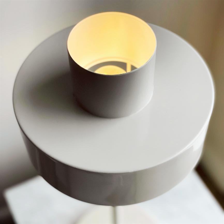 White table lamp emits warm, subdued light through a top cylindrical opening, placed on a white table top in a bright, neutral indoor space.