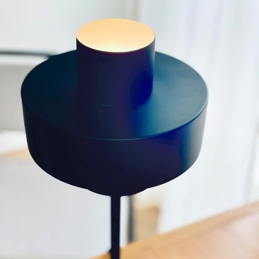 A modern black floor lamp emits warm light from a round opening in a cylindrical shade, standing by a bright window above a wooden table in a minimalist room.