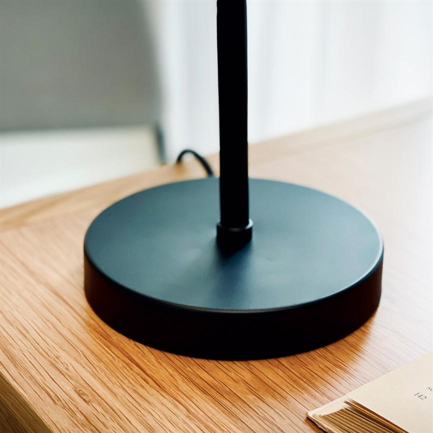 Black lamp base stands on a light wooden table, a slender rod juts up; black cord meanders behind, an open book lies in the lower right corner.