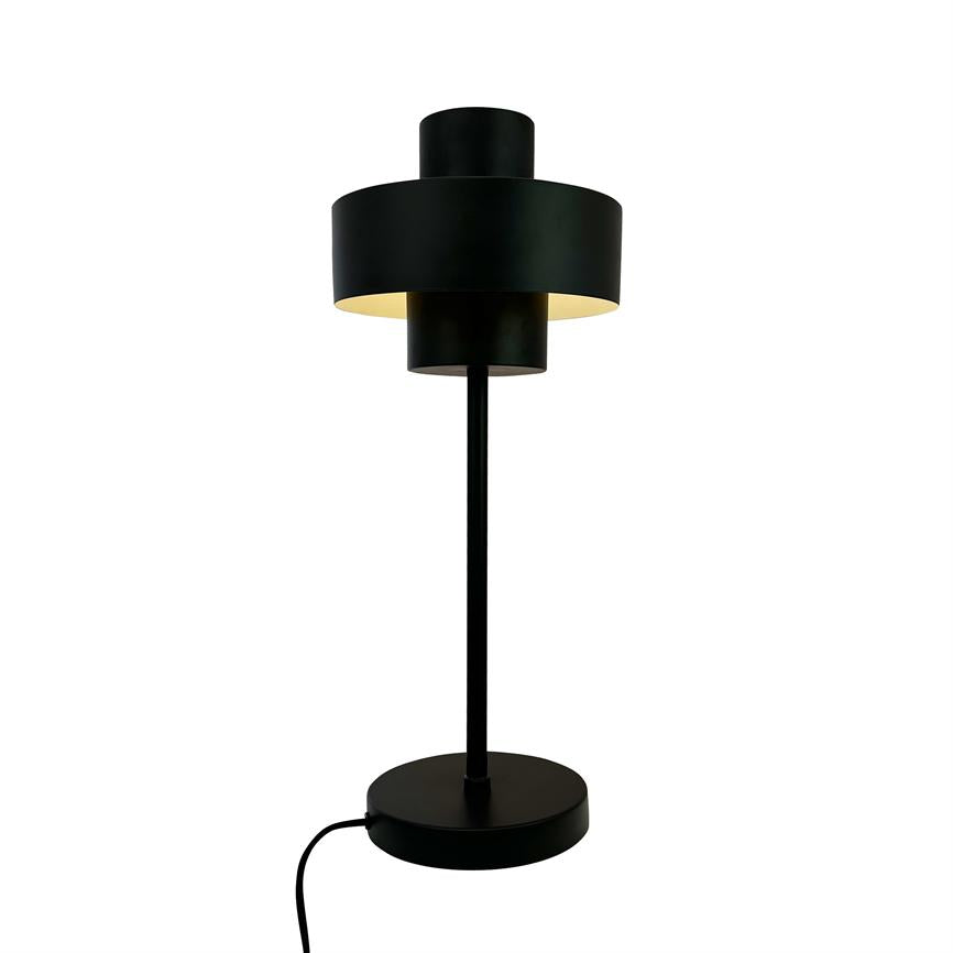 A black table lamp with overlapping cylindrical shades emits warm, downward light; it stands on a round base with visible wire against a uniform white background.