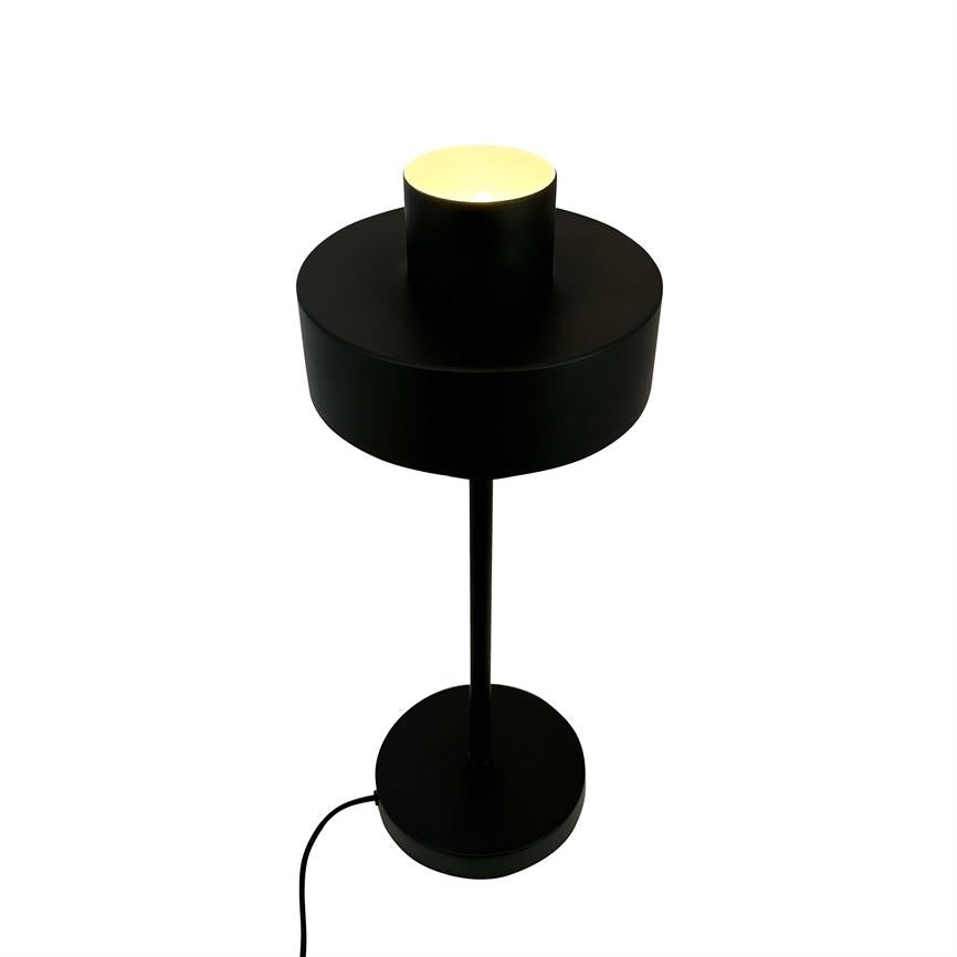 Black table lamp with cylindrical shade, emits a warm upward glow; thin rod connects screen to round foot, visible black wire, set against a uniform white background.