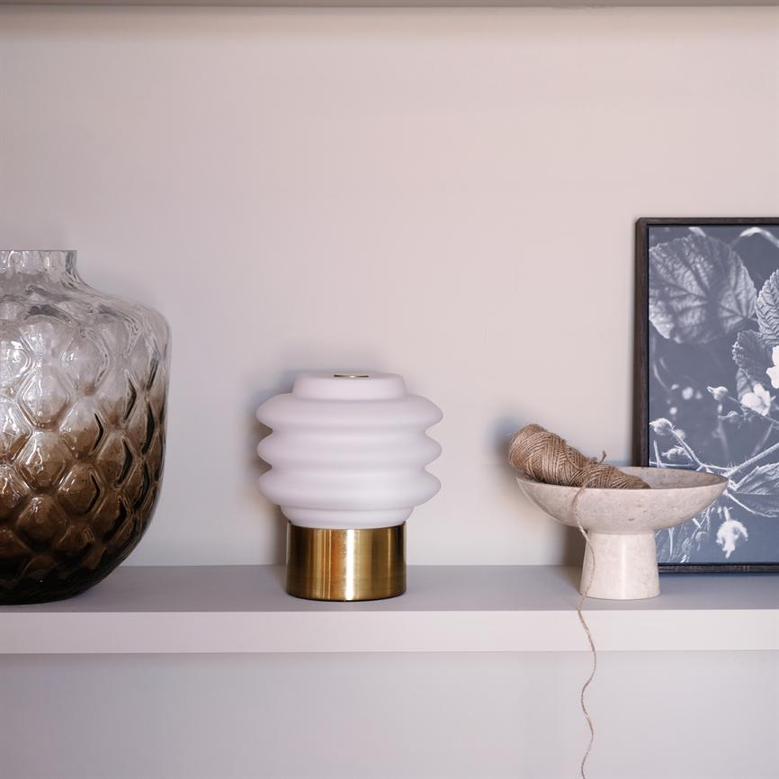 White, wavy table lamp on brass base stands on a beige shelf; on the left is a large brownish patterned glass jar, on the right a bowl with string and a black-and-white plant photo.