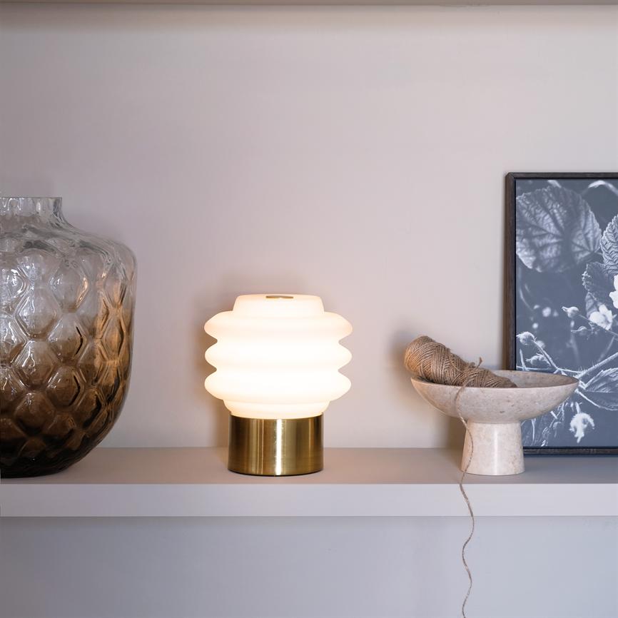 A white wavy table lamp is lit on a wall shelf between a large patterned glass vase on the left and a stone bowl with twine on the right; a framed black-and-white plant photo behind.