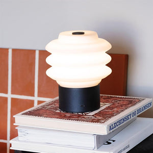 Lamp on bookshelf: A white, wavy table lamp shines softly and stands on two art books at a table with terracotta tiles in the background. Text on spine: "ALECHINSKY AND BRAMSEN".