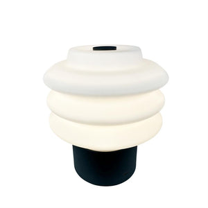 Table lamp with a white, wavy shade in three layers that shines softly; black cylinder base under the screen, against a neutral white background.